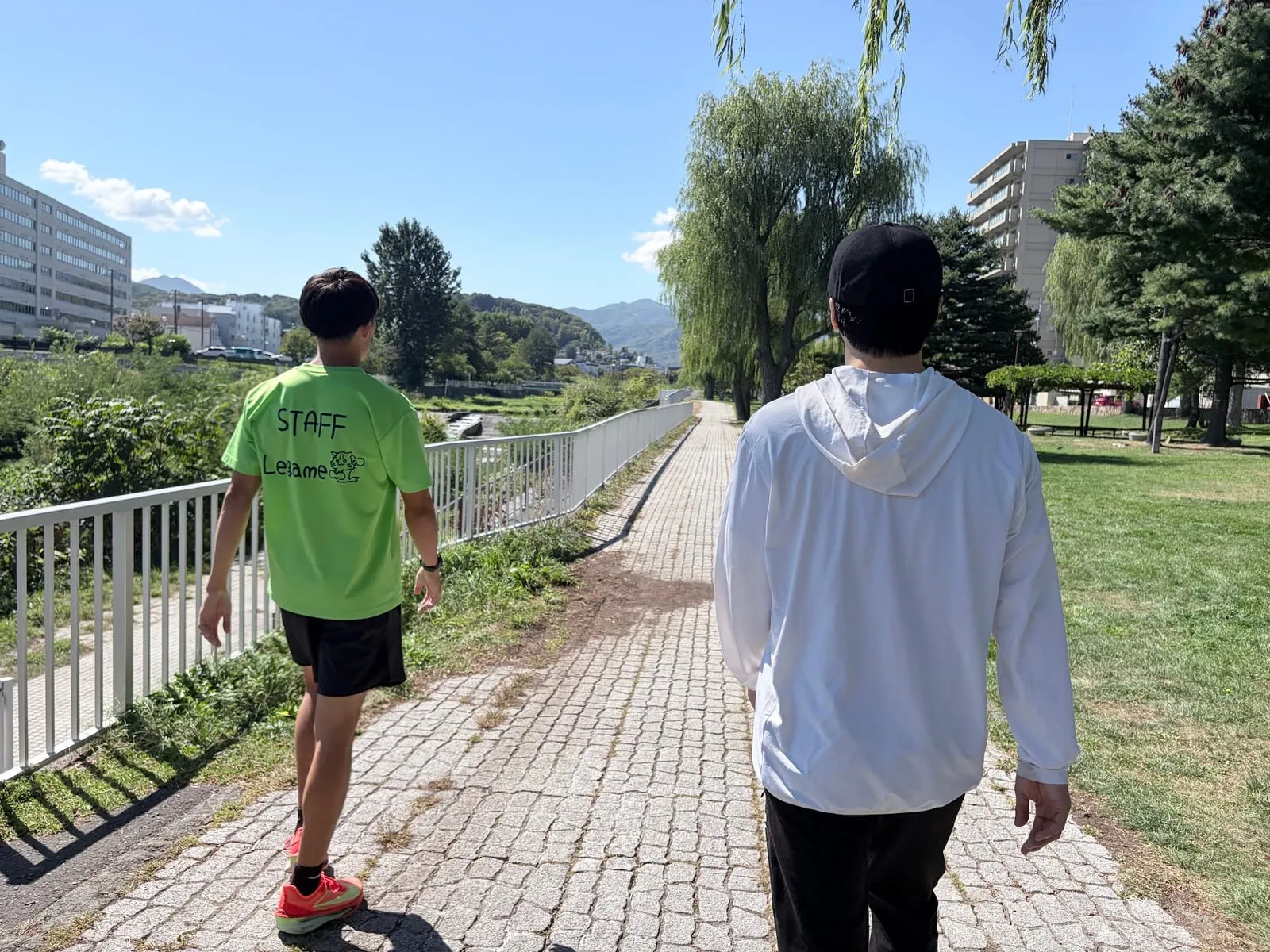 川沿いの遊歩道を歩き出すスタッフと会員(後ろ姿)